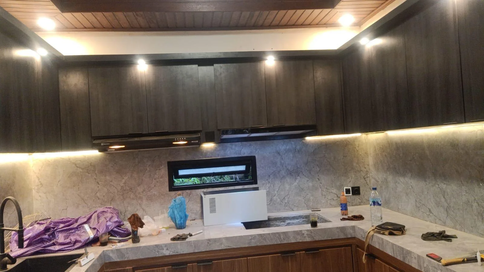 U-shaped residential kitchen with dark upper cabinets, timber-look lower cabinets, stone-look surfaces, task lighting, and a timber ceiling; installation items remain on the counter.