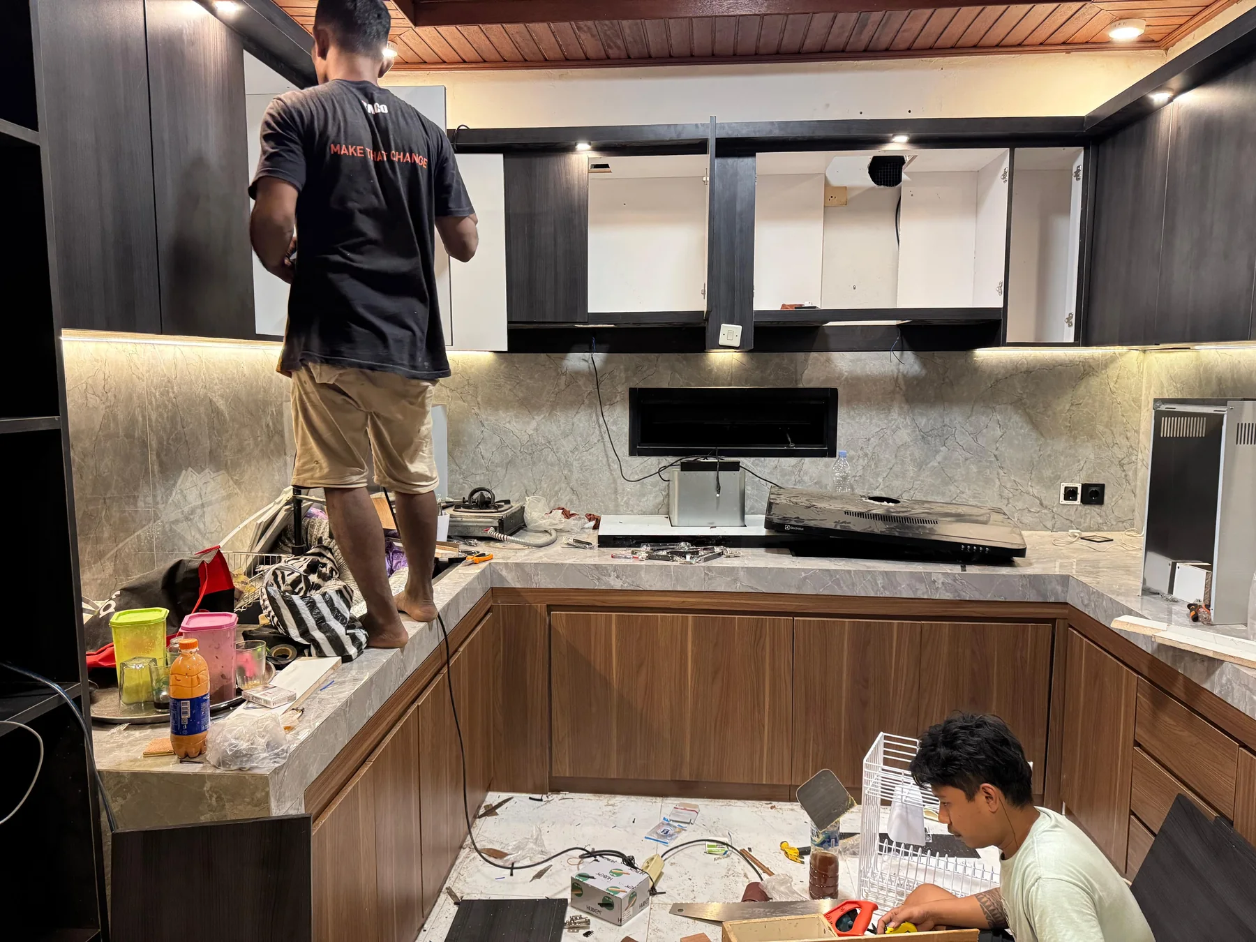 Kitchen renovation in progress with open upper cabinets, installed lower cabinetry, task lighting, and workers fitting custom elements.