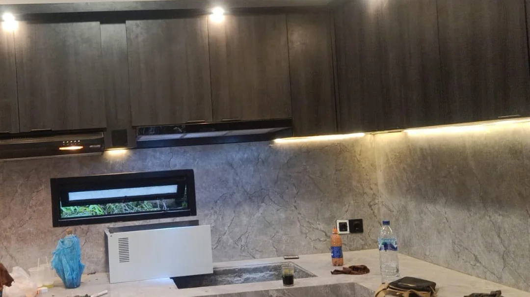 Detail of dark upper cabinetry, integrated task lighting, stone-look backsplash, and the small horizontal kitchen window.